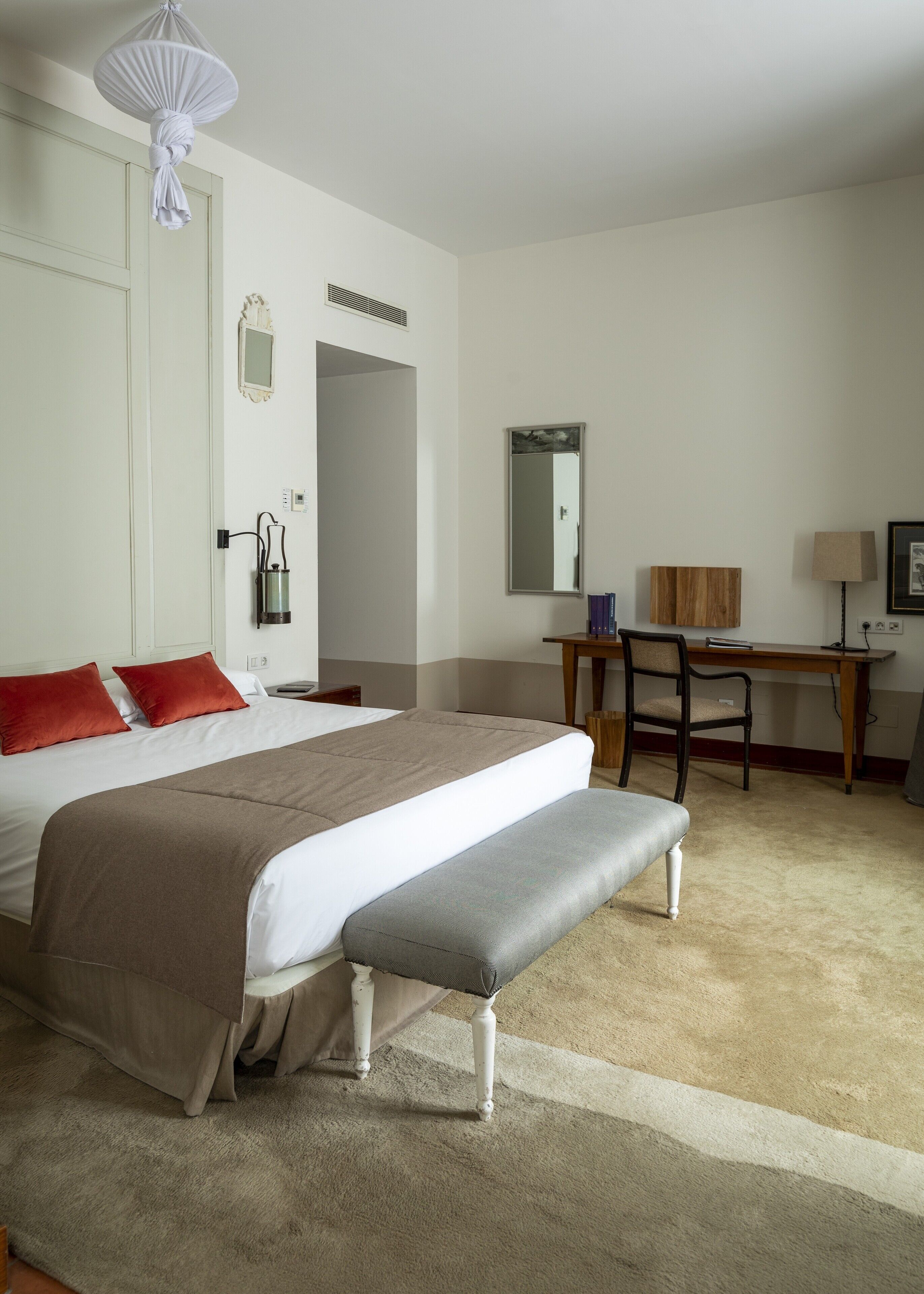 Une chambre d’hôtel comprenant un lit, un bureau, une chaise, un banc, un miroir et un luminaire suspendu.