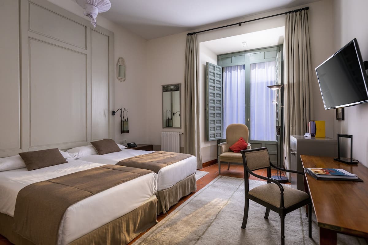 A hotel room with two beds, a desk, a chair, a TV, and a view of the outdoors.