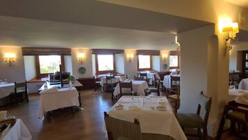 A spacious dining area with multiple tables and chairs, well-lit by wall-mounted lamps, and large windows providing natural light.