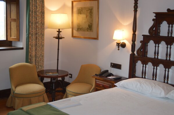 A hotel room with a bed, two chairs, a small table, a lamp, a painting, and a telephone.