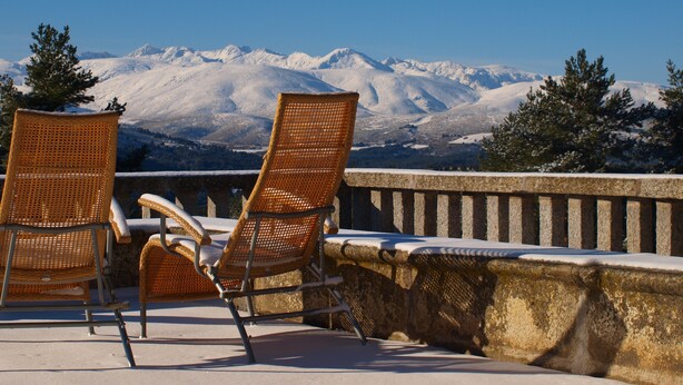 Two wicker chairs with metal frames and a stone balustrade, overlooking a snowy mountain range.