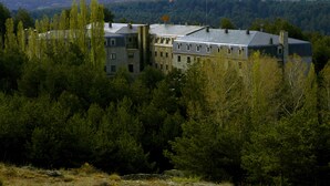 A large, historic building with multiple windows and a flat roof, surrounded by dense forest and mountains in the background.