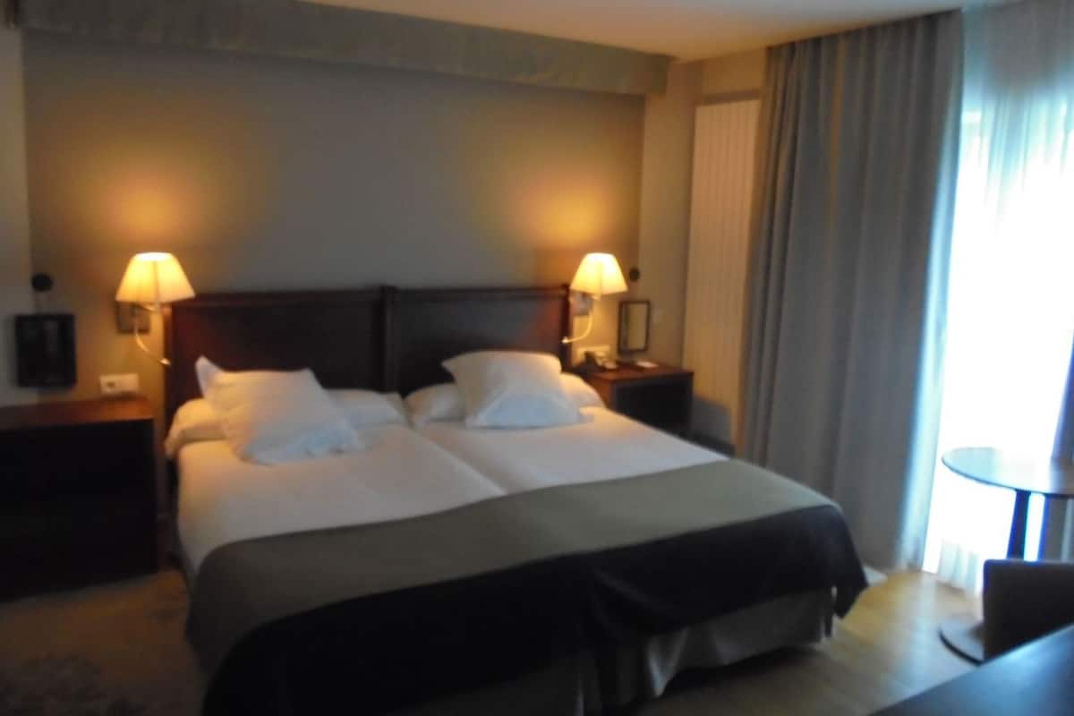 A hotel room with a double bed, two bedside lamps, a nightstand, a small table, and a view of the outdoors through a window with curtains.