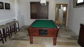 A pool table in a room with stone flooring, wooden stools, and a mirror on the wall.