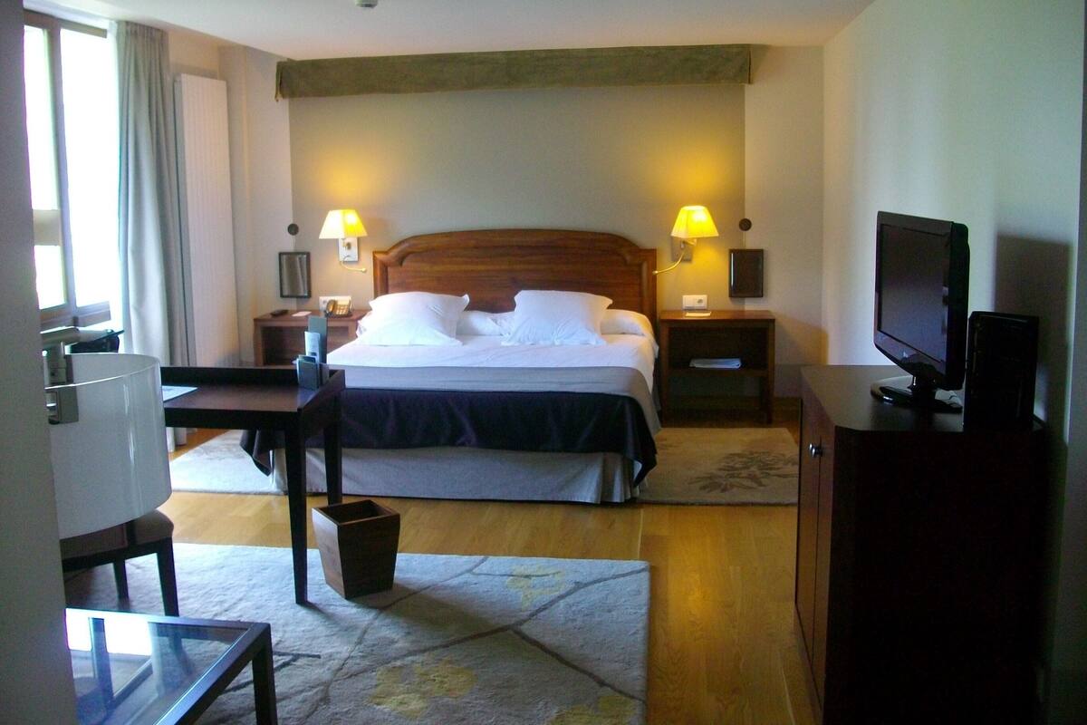 A hotel room with a bed, two bedside lamps, a television, a desk, and a chair.