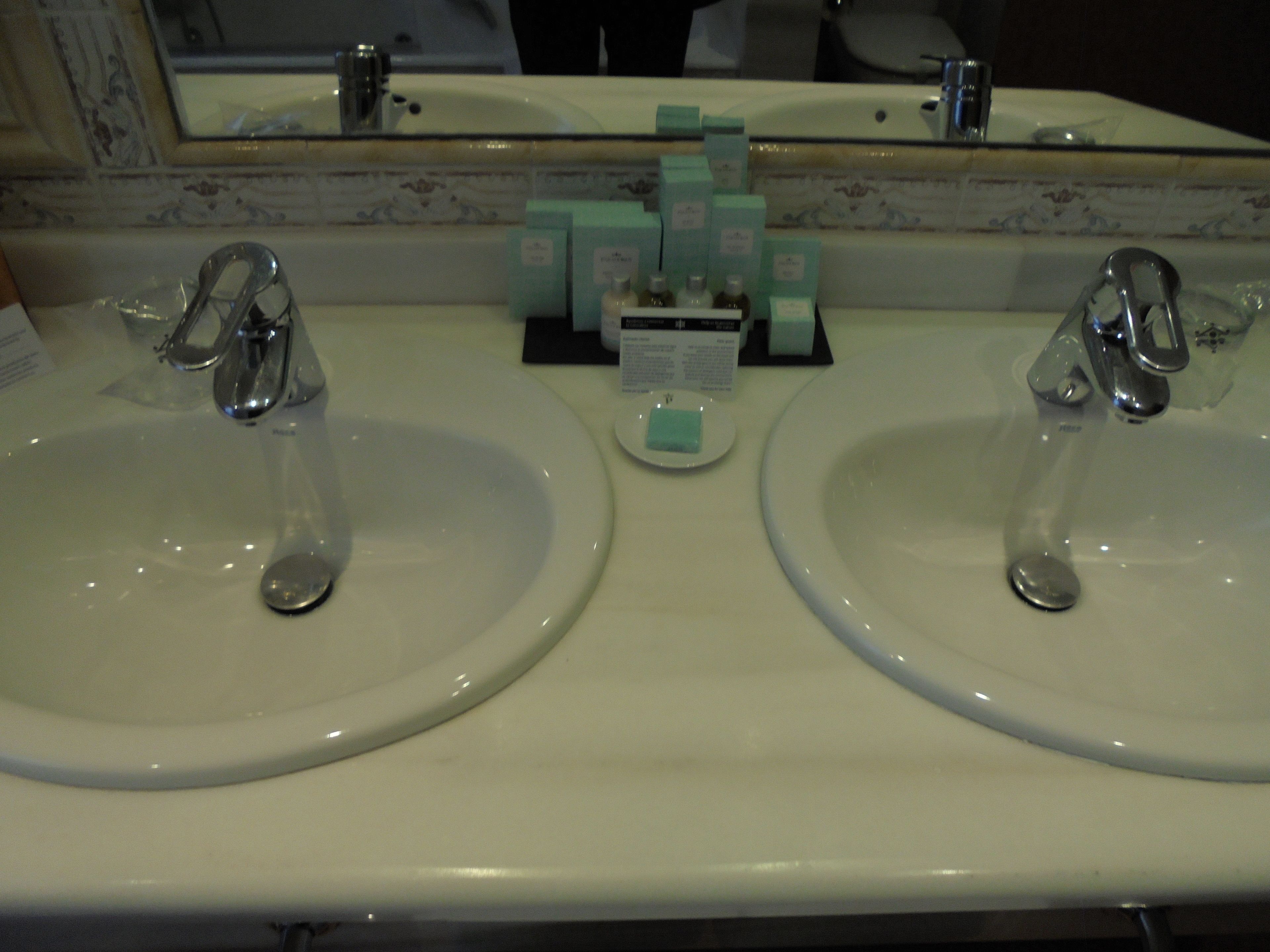 Un baño con dos lavamanos, cada uno con un grifo, y un espejo encima. Hay artículos de aseo en la encimera.