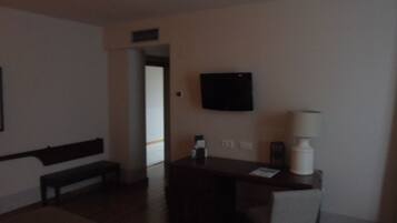 A dimly lit hotel room with a wooden bench, a small table, a chair, and a flat-screen TV mounted on the wall.