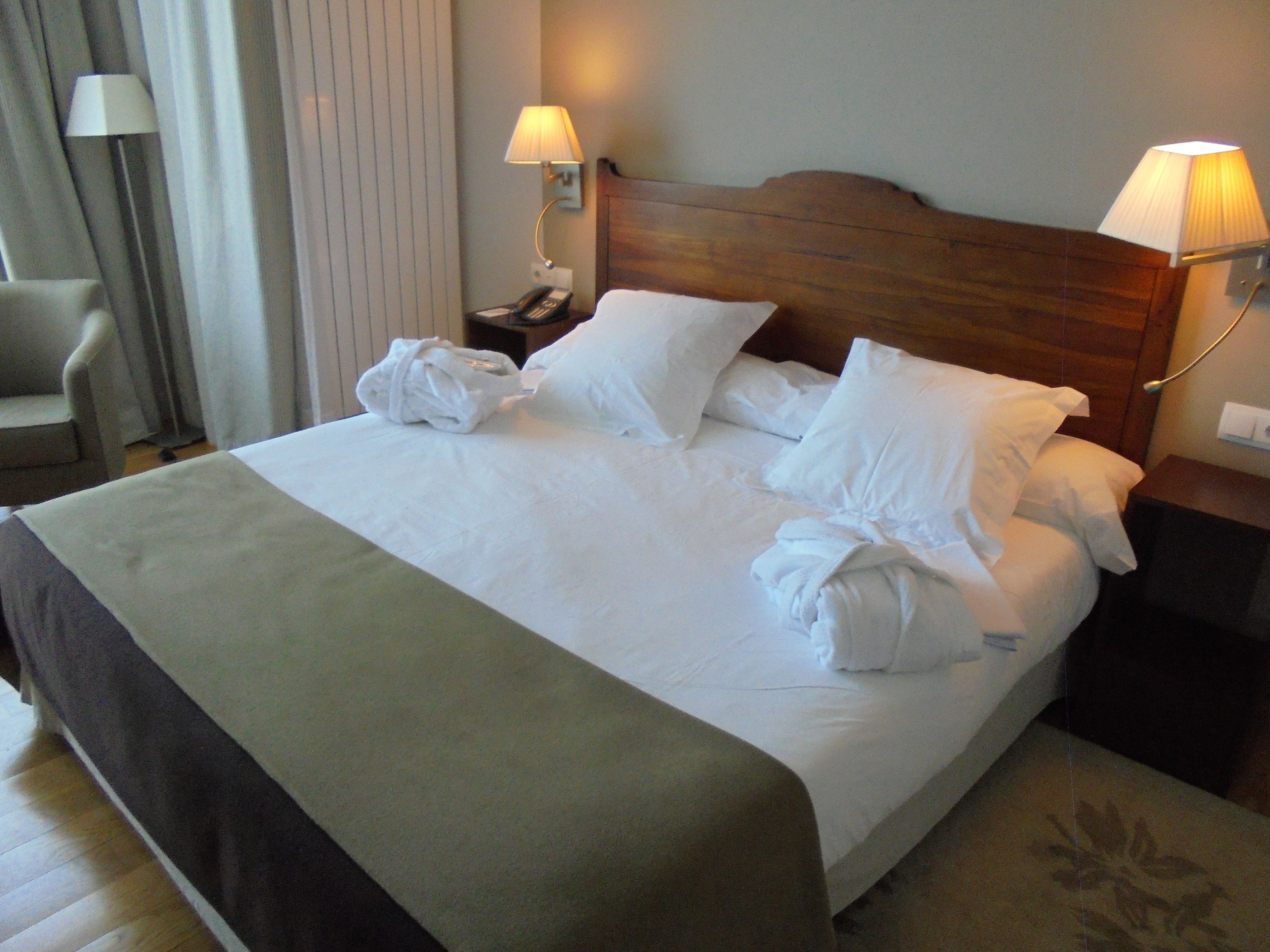 Una cama bien hecha con sábanas blancas, dos lámparas de noche, un cabecero de madera y una silla con lámpara.