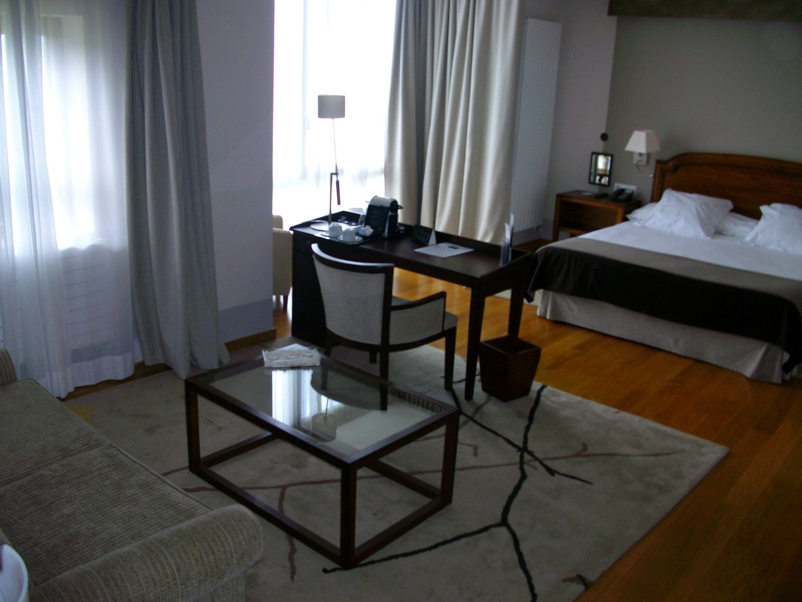 A hotel room with a bed, a desk with a chair, a glass-top table, a nightstand with a lamp, and a window with curtains.