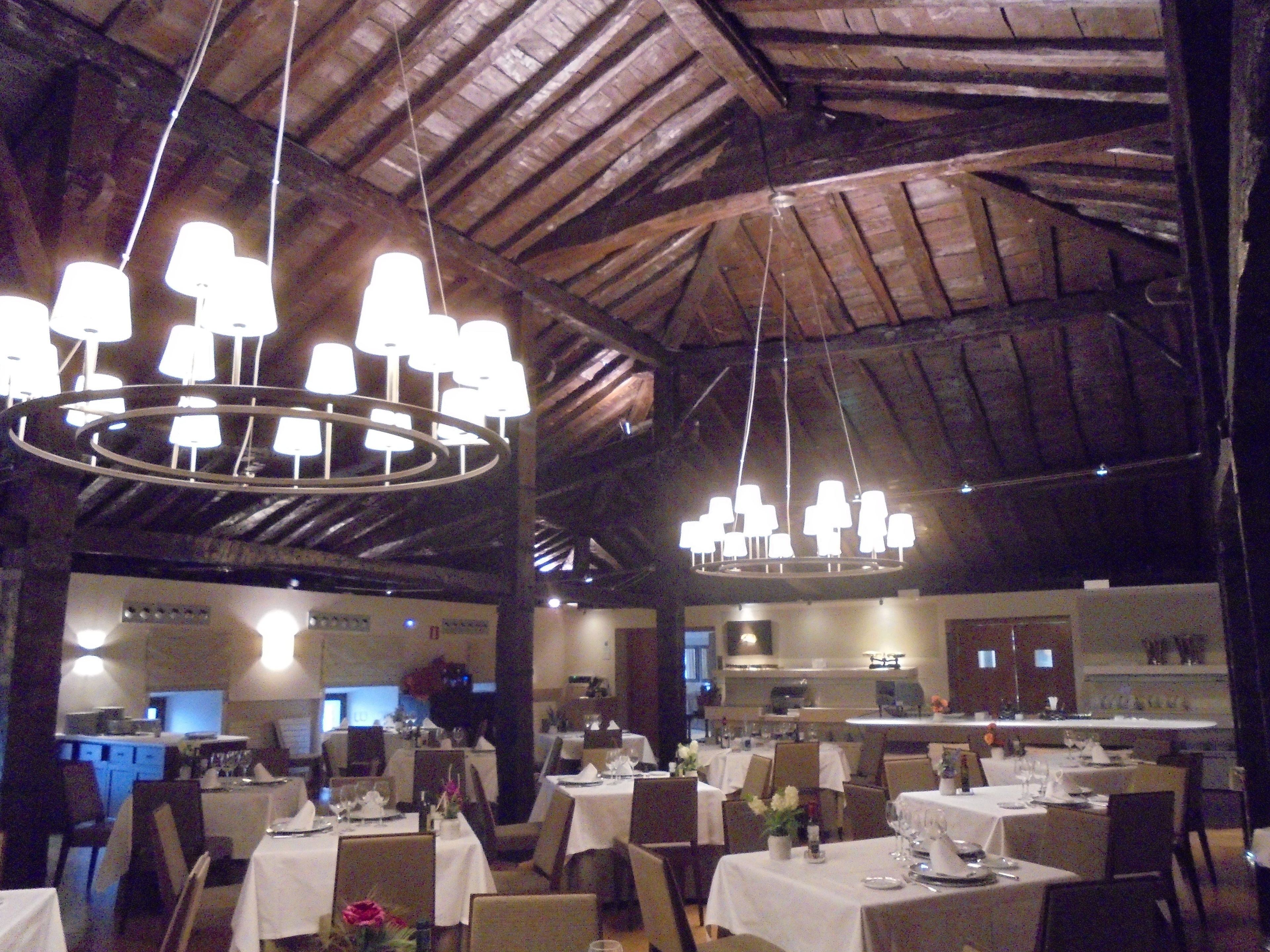A restaurant with multiple tables, a kitchen in the background, and a wooden ceiling.