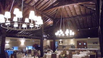 A restaurant with multiple tables, a kitchen in the background, and a wooden ceiling.