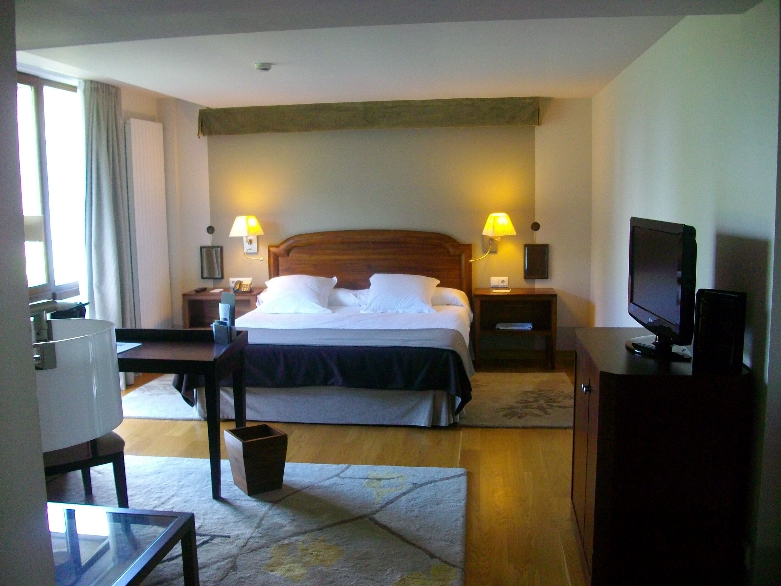 Una habitación de hotel con una cama, dos lámparas de noche, un televisor, un escritorio y una silla.