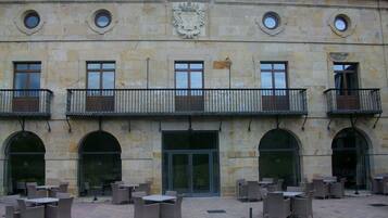 Un edificio con fachada de piedra, múltiples ventanas y una entrada central. Hay zonas de estar al aire libre con mesas y sillas.