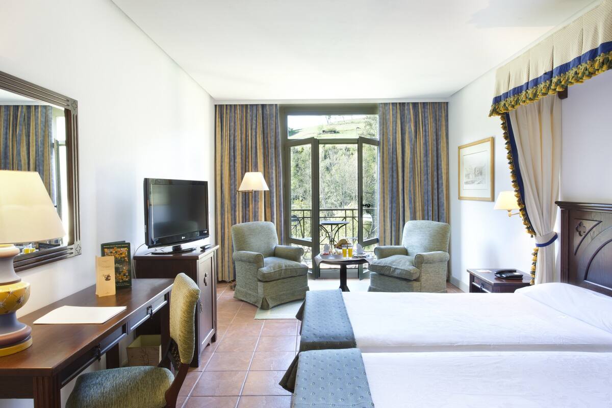 A hotel room with a bed, two armchairs, a desk with a computer, a TV, and a window with curtains.