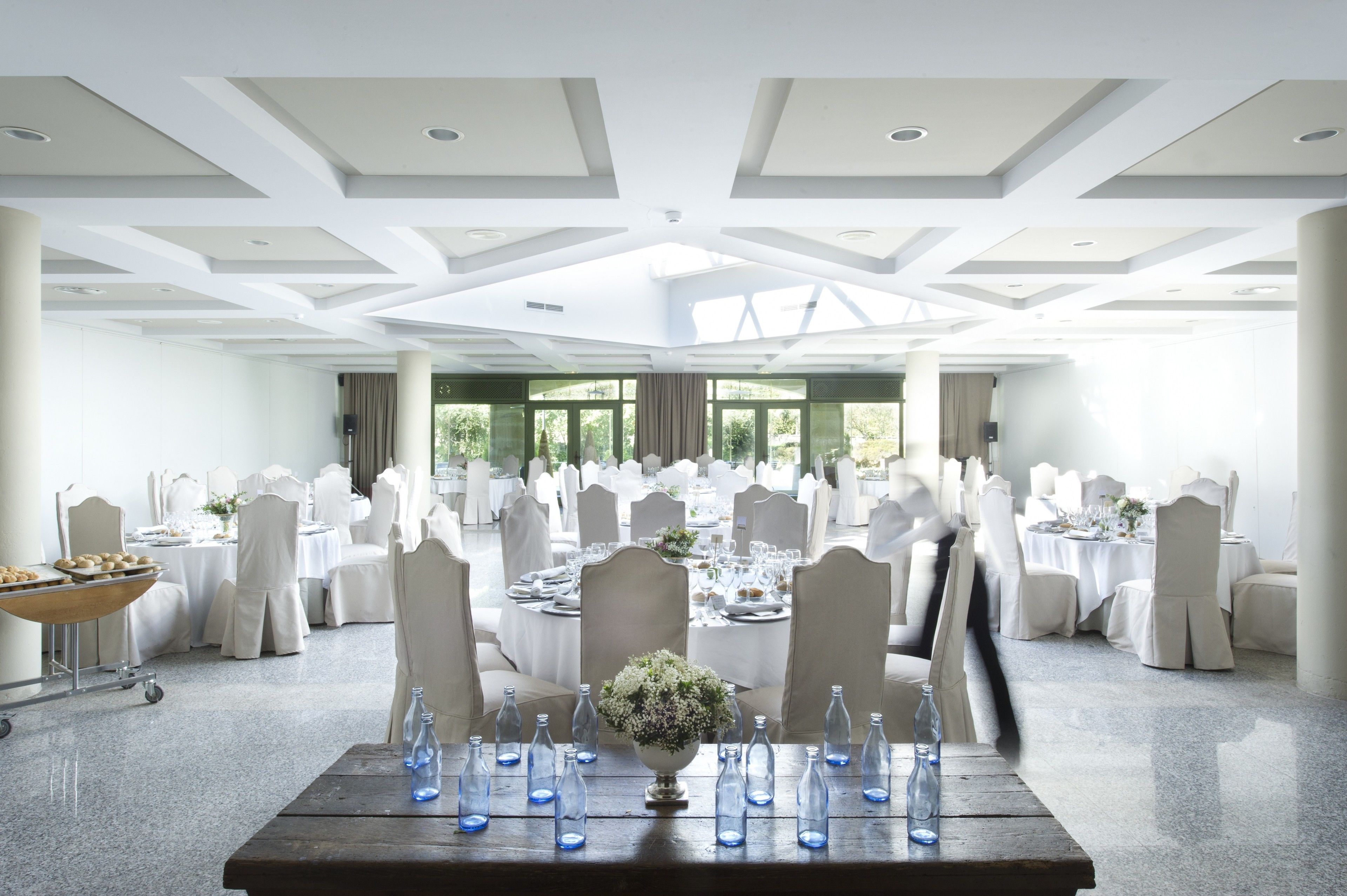 Un amplio salón de banquetes con mesas y sillas blancas, un arreglo floral central y grandes ventanales que dejan entrar luz natural.