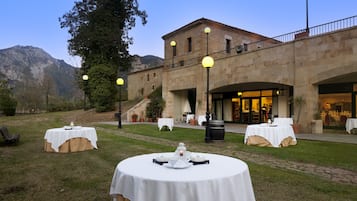 Un'area pranzo all'aperto con tavoli rotondi apparecchiati per un pasto, un edificio in pietra con un balcone sullo sfondo e una catena montuosa all'orizzonte.
