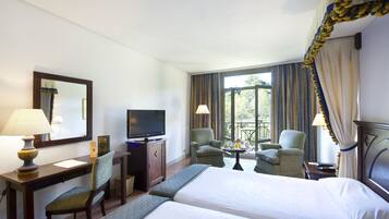 A hotel room with two beds, a desk with a TV, a chair, a small table with a lamp, and a view of the outdoors through large windows.
