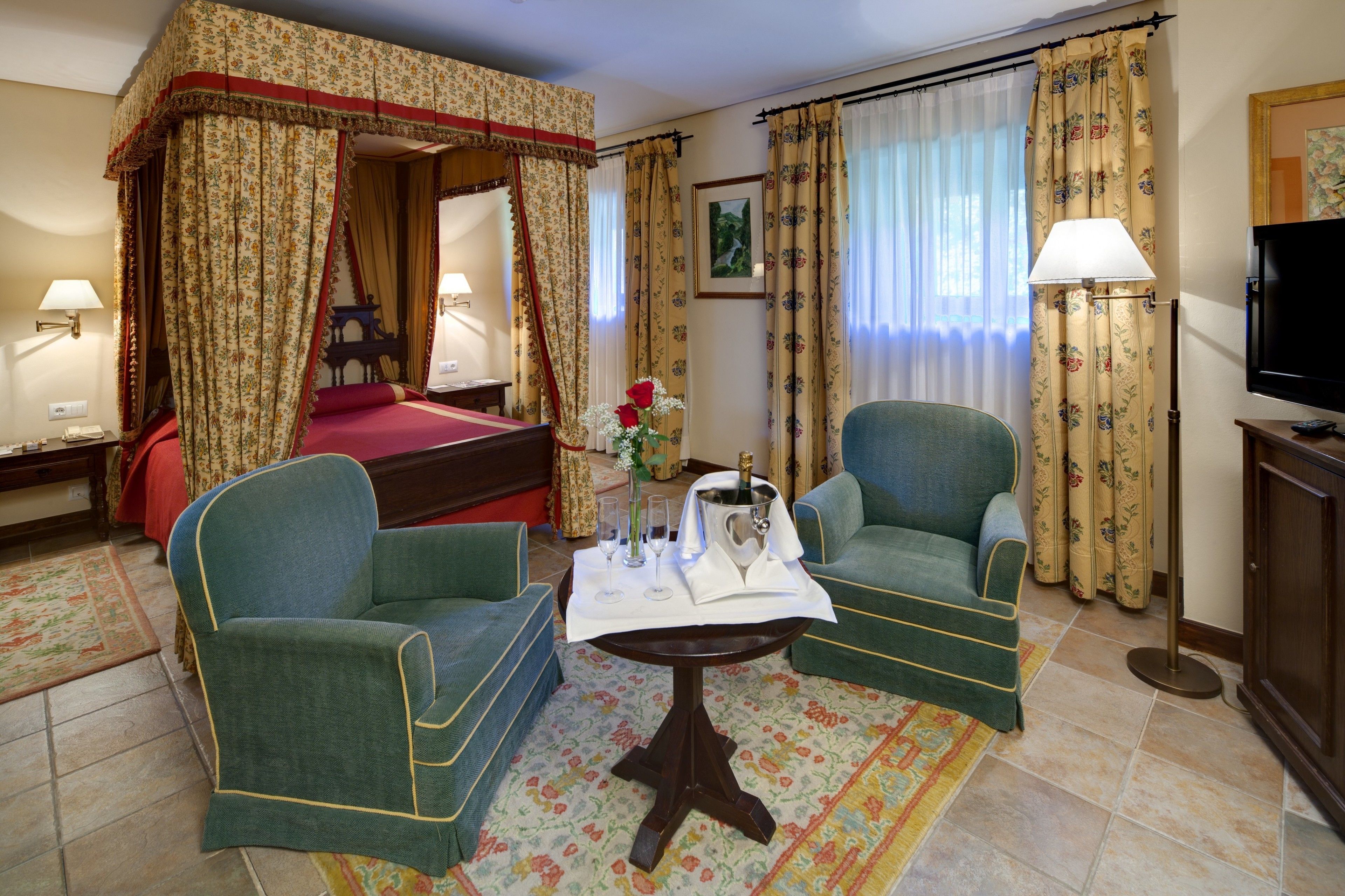 Una habitación con una cama con dosel, dos sillones, una mesita con un florero, un televisor y una lámpara.