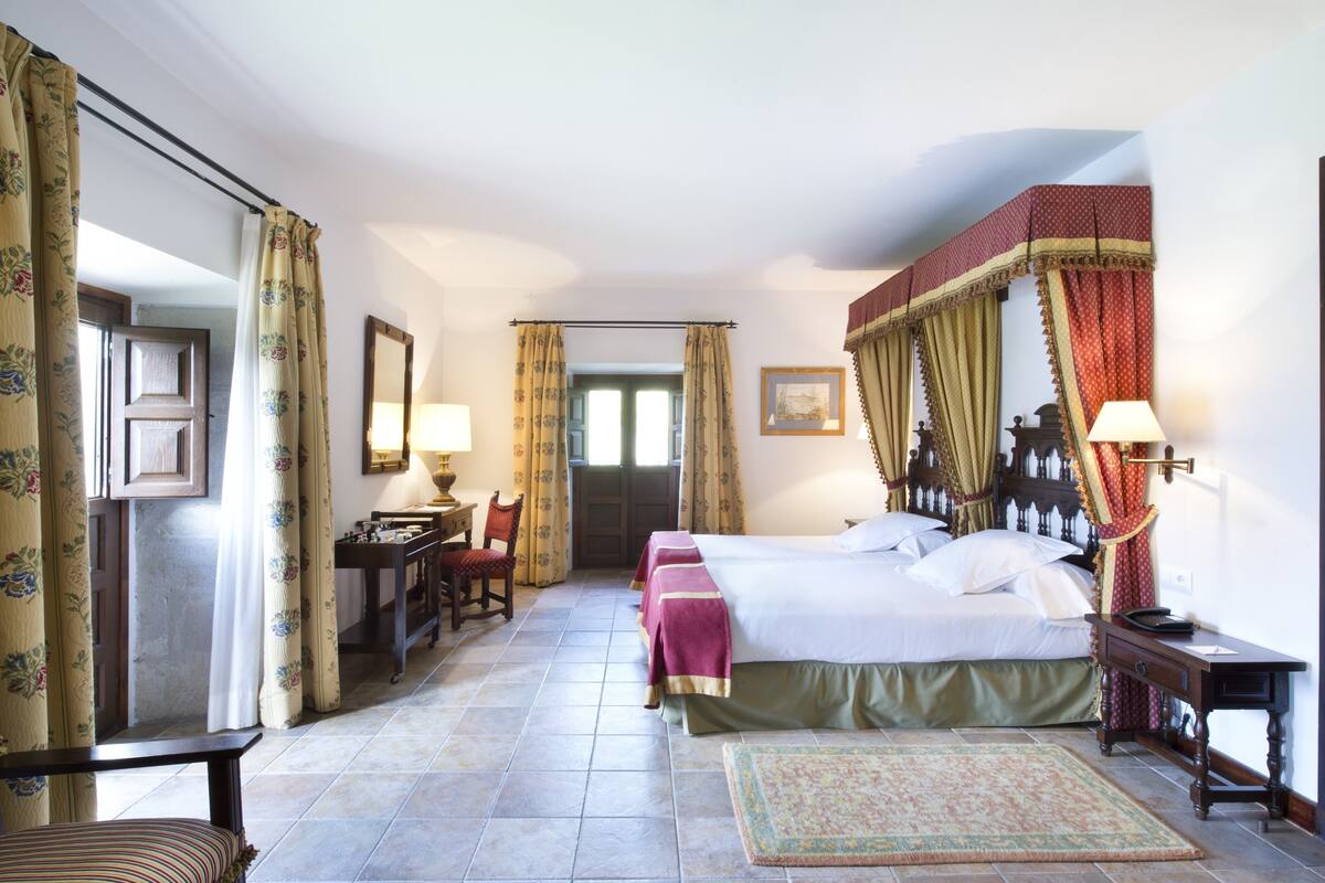 A hotel room with a large bed, a desk with a chair, a desk lamp, and a nightstand with a lamp. There is a door leading to another room and a window with curtains.