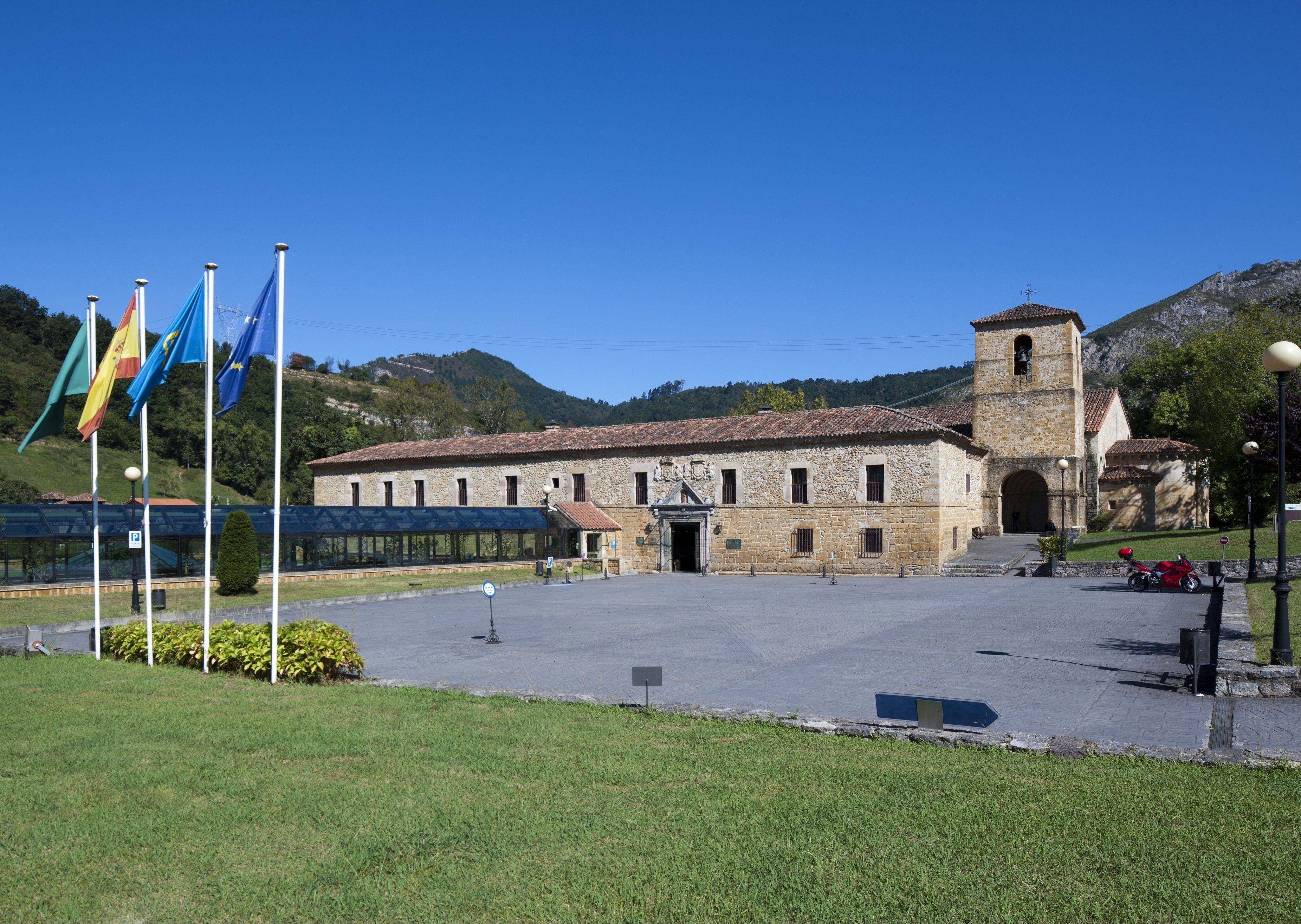 Un edificio de piedra histórico con un campanario, rodeado de una zona pavimentada con banderas y un cielo despejado.
