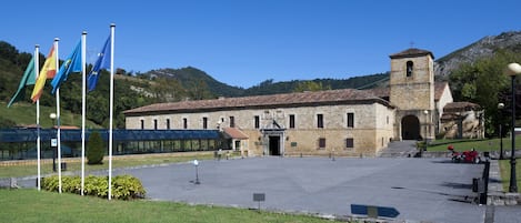 Un edificio storico in pietra con un campanile, circondato da un’area pavimentata con bandiere e un cielo sereno.