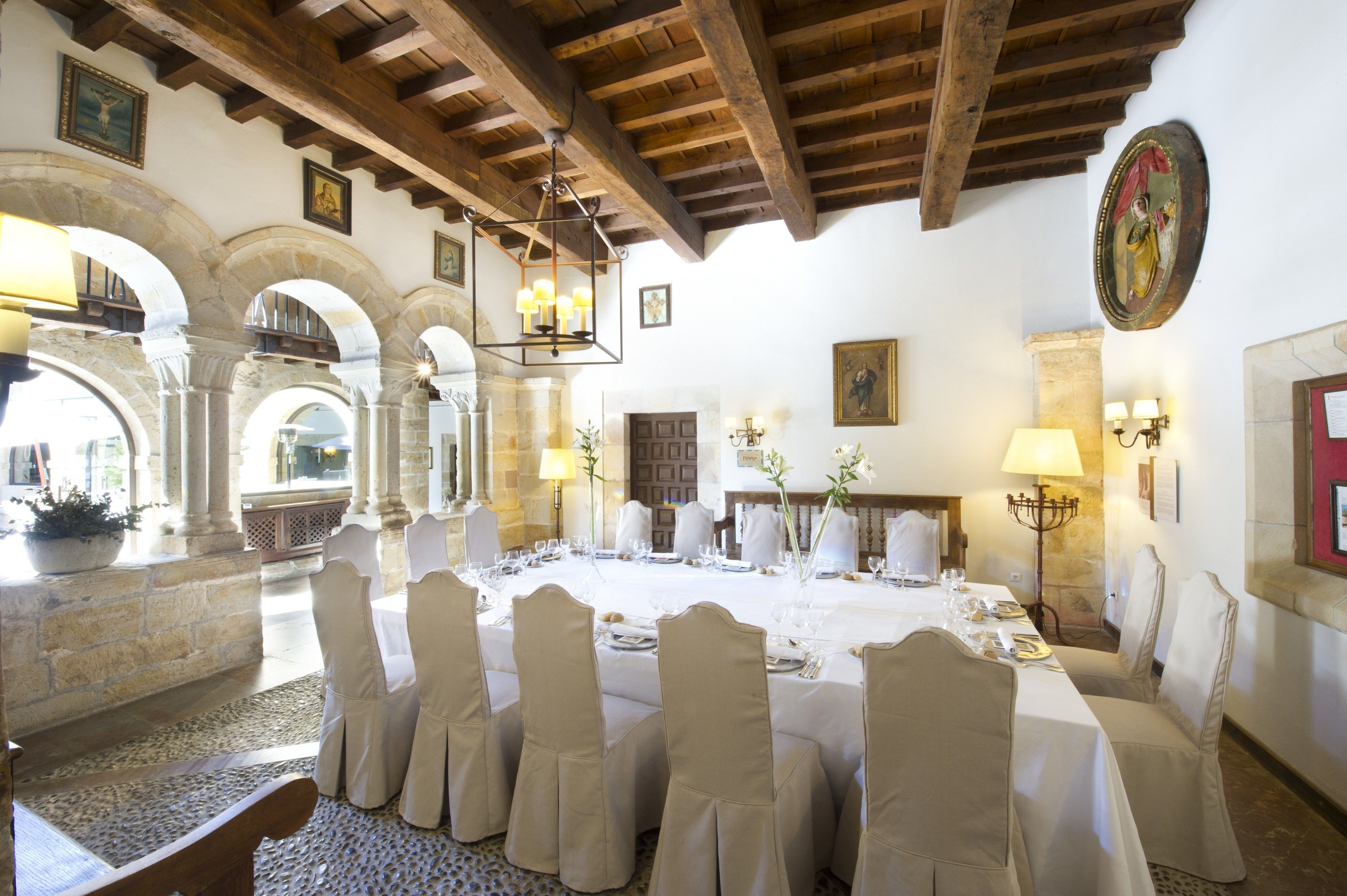 A dining area with a long table set for a meal, surrounded by chairs, in a room with stone arches and wooden beams.