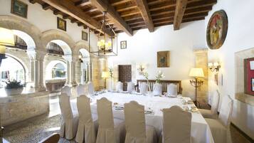 A dining area with a long table set for a meal, surrounded by chairs, in a room with stone arches and wooden beams.