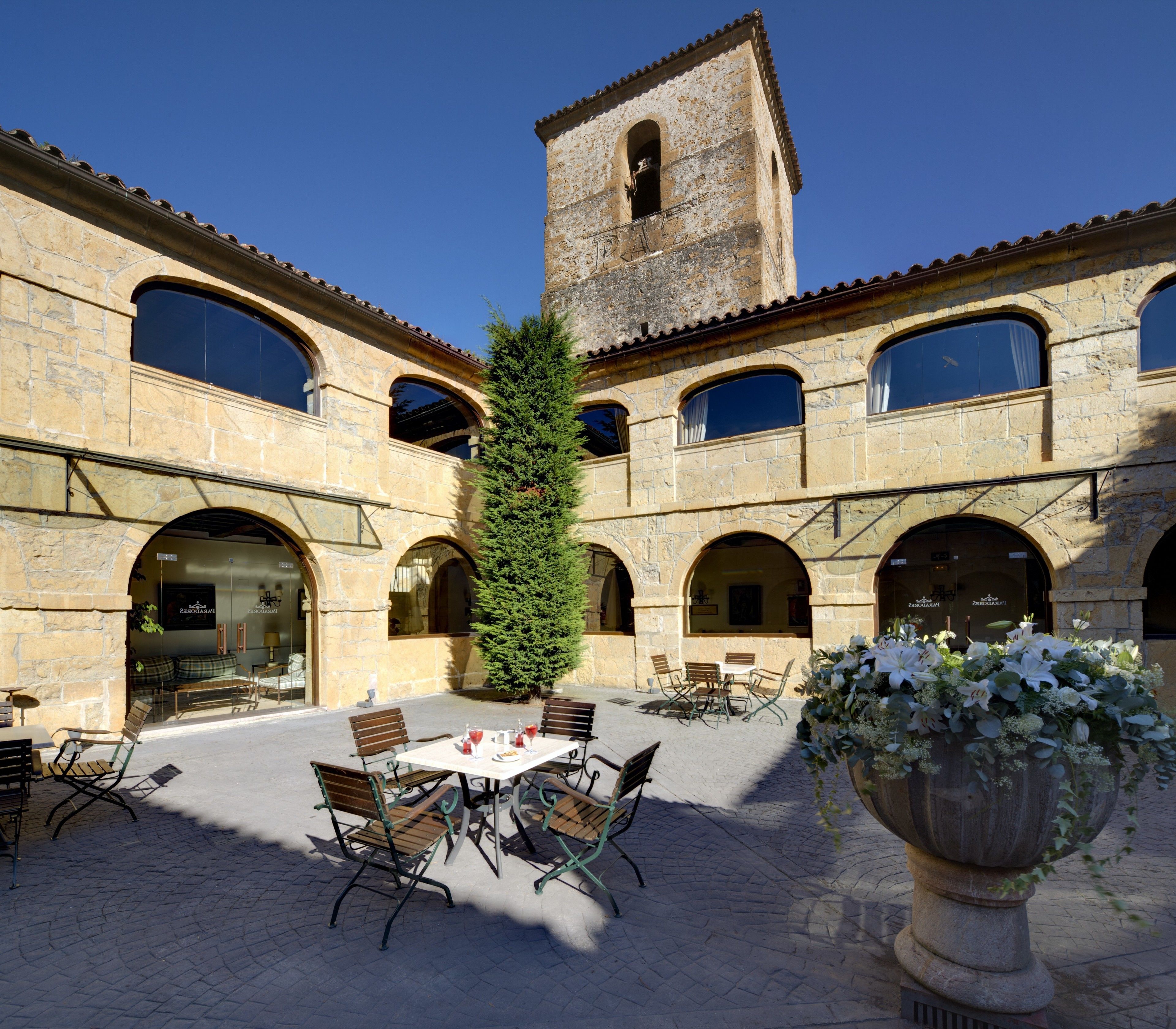 A courtyard with stone buildings, a bell tower, outdoor seating, and a large planter with flowers.