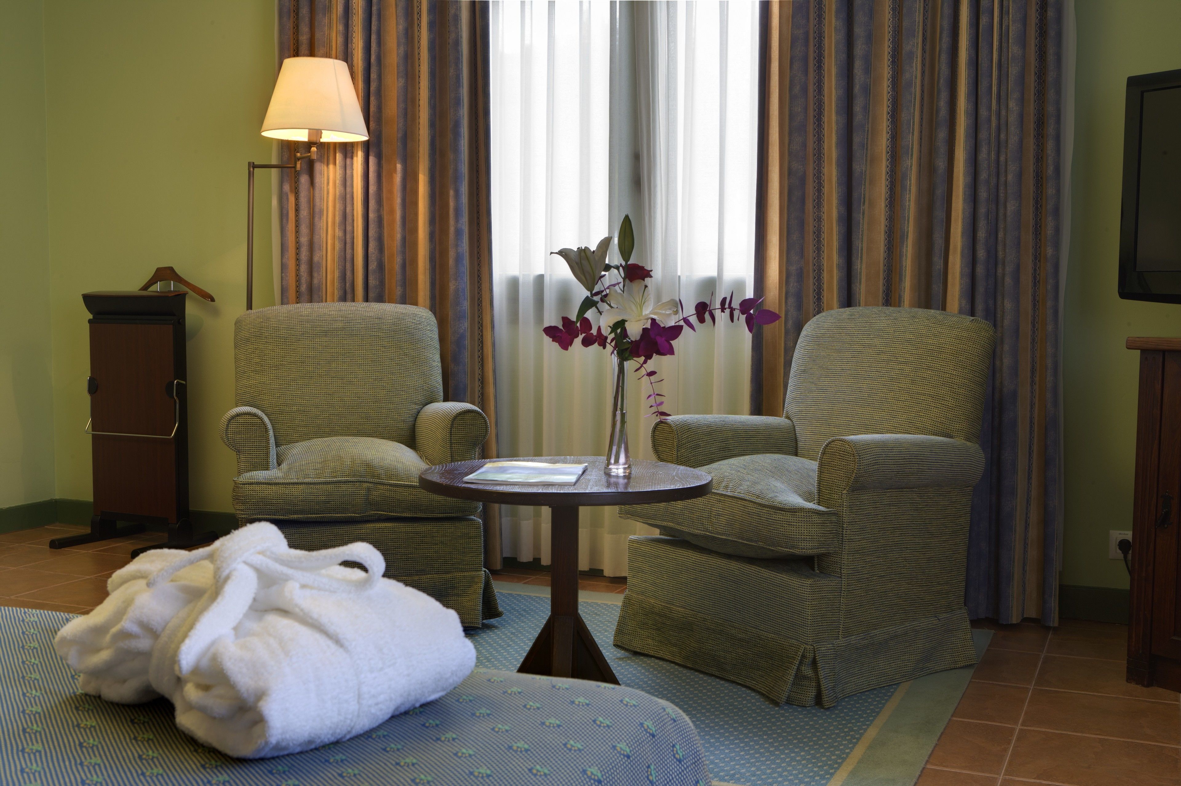 Una habitación de hotel con dos sillones verdes tapizados, una mesita redonda con una lámpara, un jarrón con flores y un par de toallas blancas en el suelo.