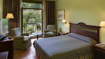 Una camera d'hotel con un letto grande, due poltrone, un tavolino e un balcone con vista sul verde.