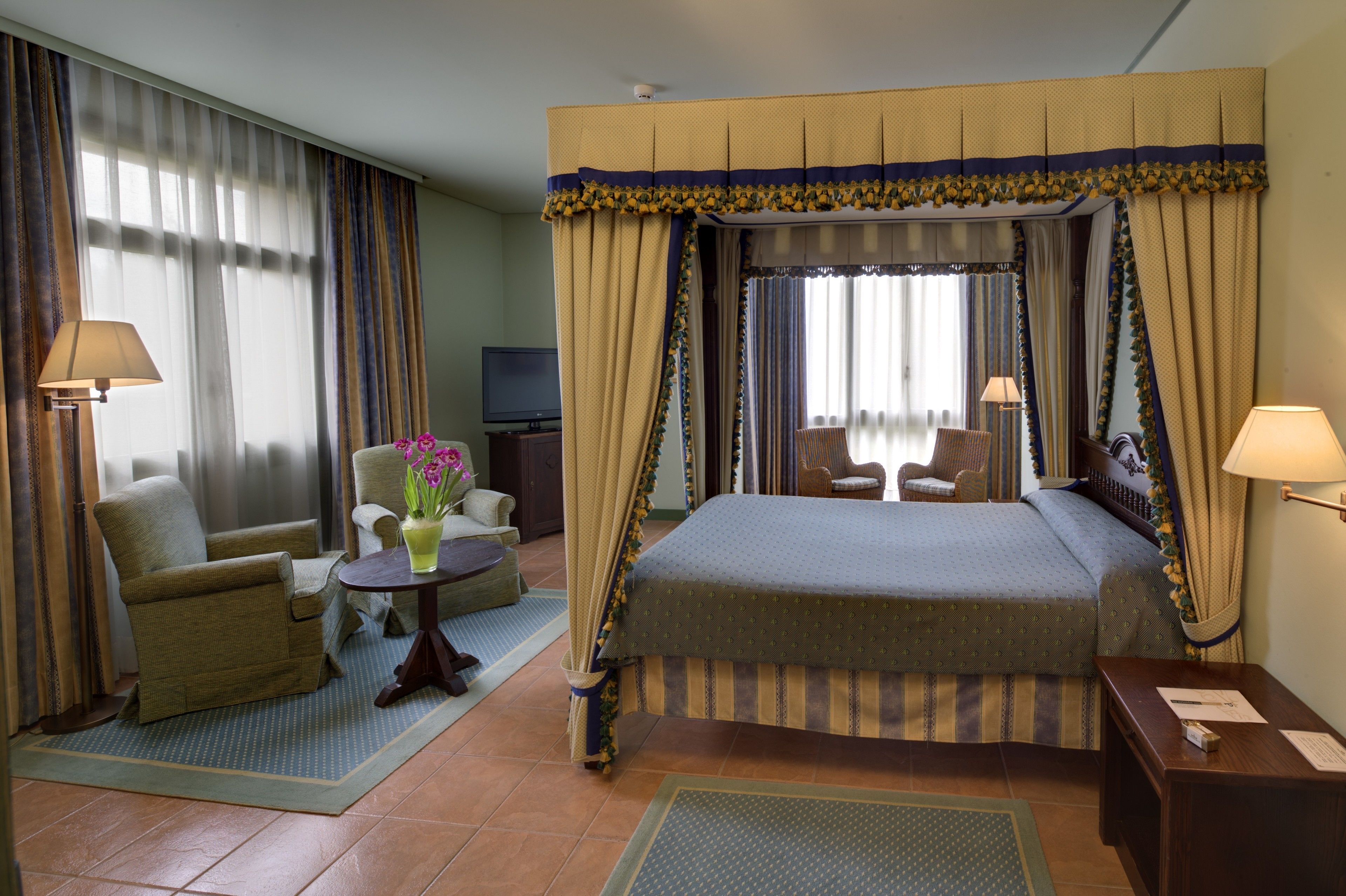 Habitación de hotel con cama con dosel, una zona de estar con una silla y una mesita, un televisor y una lámpara.
