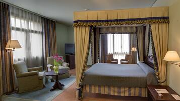 Habitación de hotel con cama con dosel, una zona de estar con una silla y una mesita, un televisor y una lámpara.