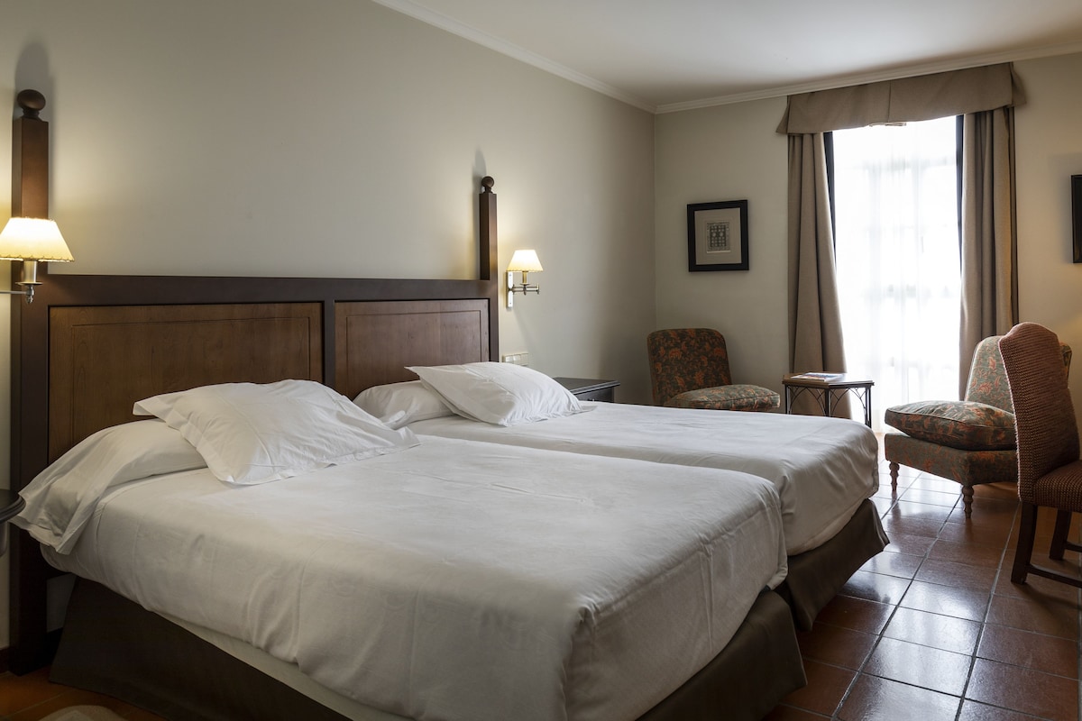 A hotel room with two beds, a chair, a small table, and a window with curtains.