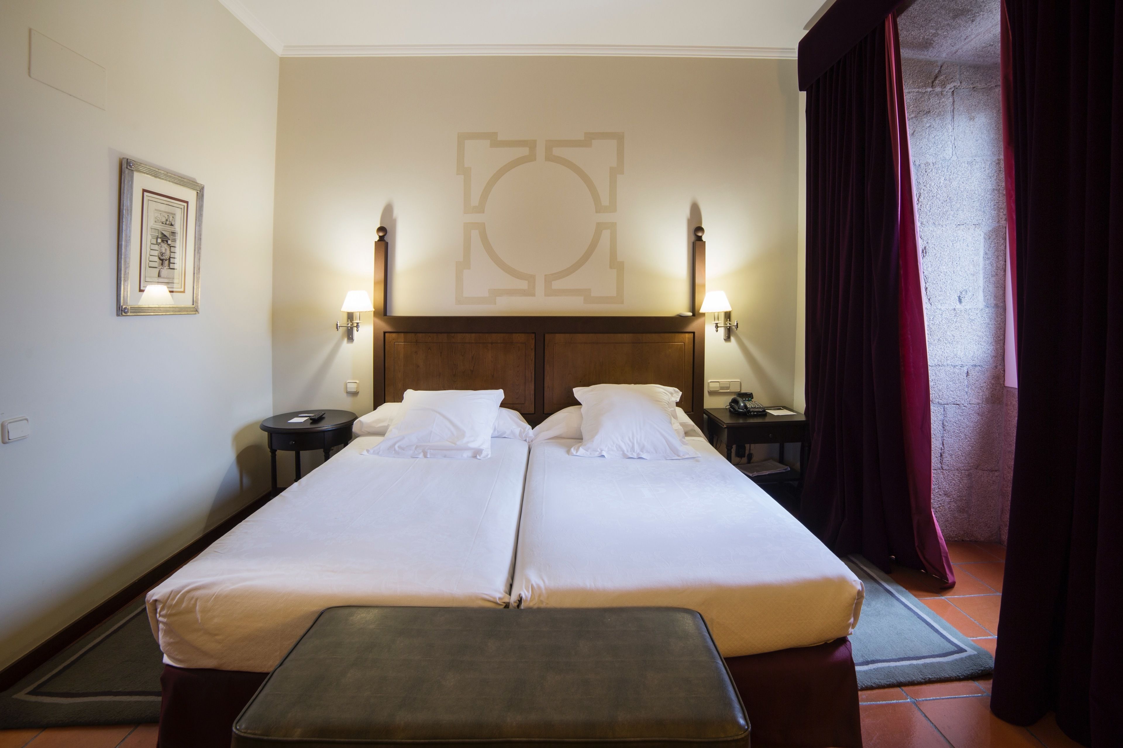 A double bed with a wooden headboard, two bedside tables with lamps, a framed picture on the wall, and a red curtain partially drawn.