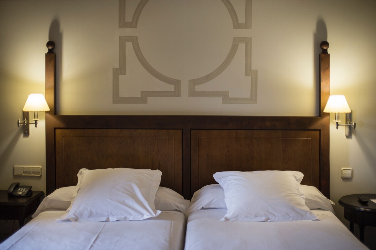 A hotel room with two beds, wooden headboard, wall sconces, a phone on the nightstand, and a small table with a remote control.