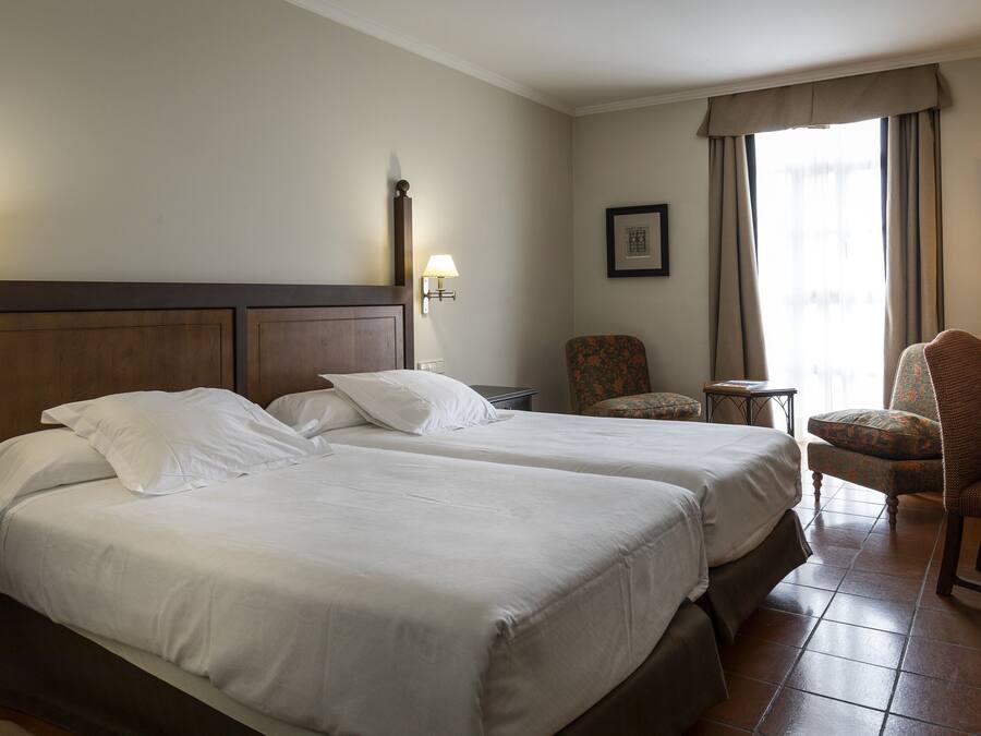 Habitación de hotel con dos camas, una silla, una mesita y una ventana con cortinas.