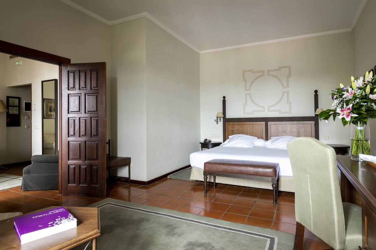 A hotel room with a bed, a desk, a chair, a bedside table, a lamp, and a vase with flowers.