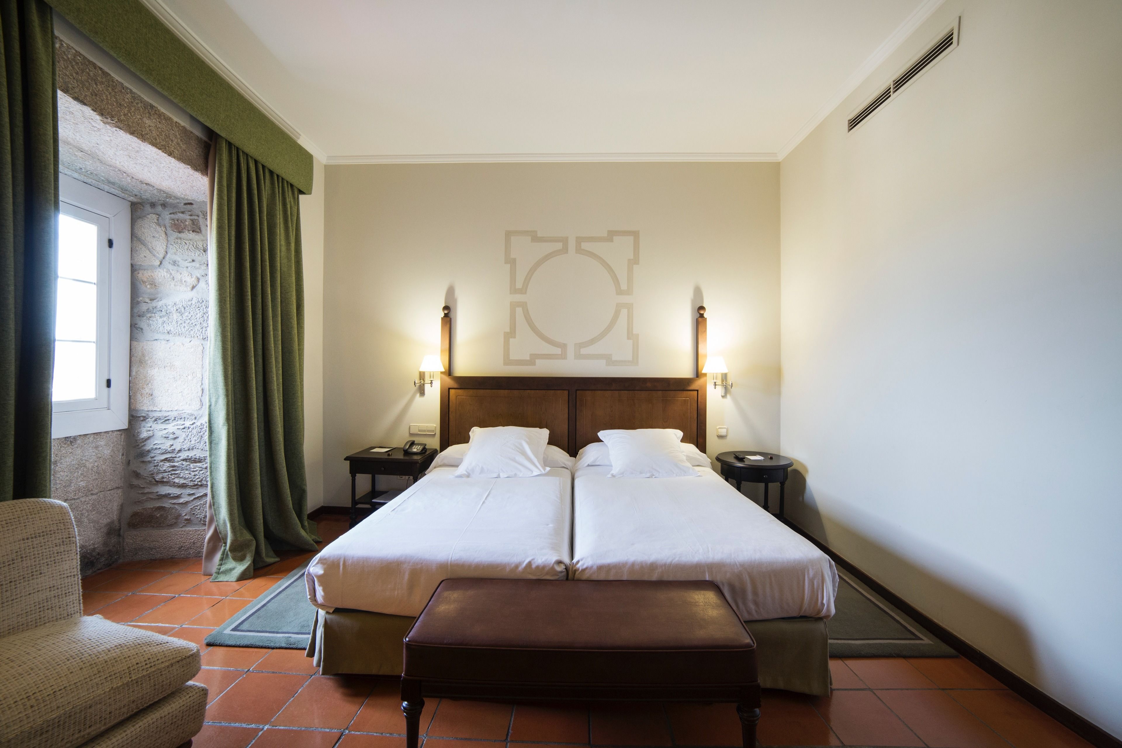 Un lit double avec deux tables de chevet, un banc, une chaise et un mur en pierre.