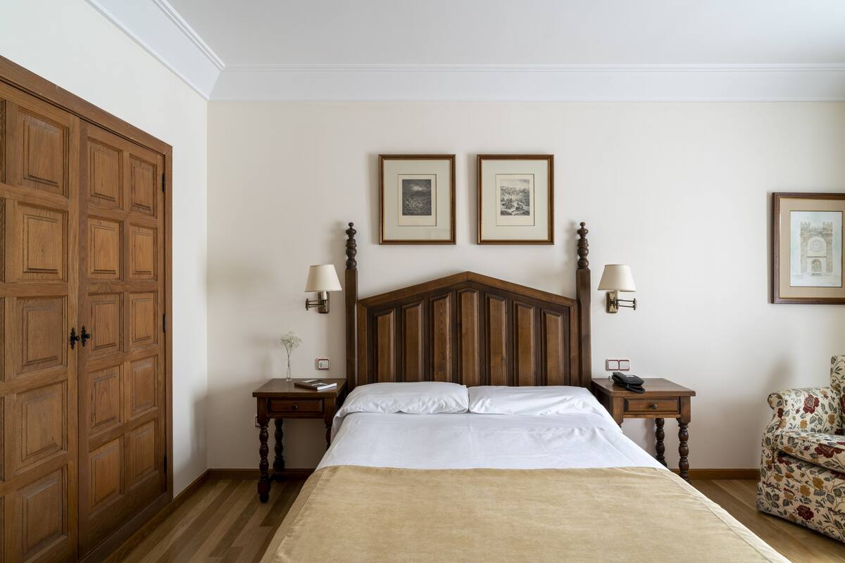 Quarto com cabeceira de madeira, duas mesas de cabeceira com luminárias, cama com lençóis brancos e duas molduras na parede.
