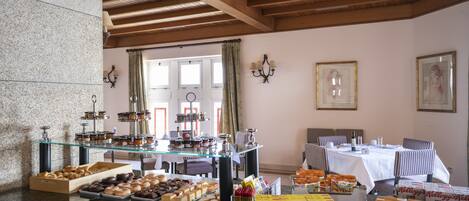 A breakfast buffet with various pastries and boxed cereals, a seating area with tables and chairs, and a view of the outdoors through large windows.