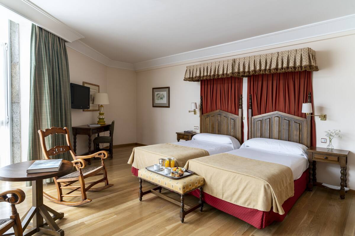 Quarto de hotel com duas camas, uma cadeira de balanço de madeira, uma mesa pequena com uma bandeja de itens de café da manhã e uma televisão sobre um suporte.