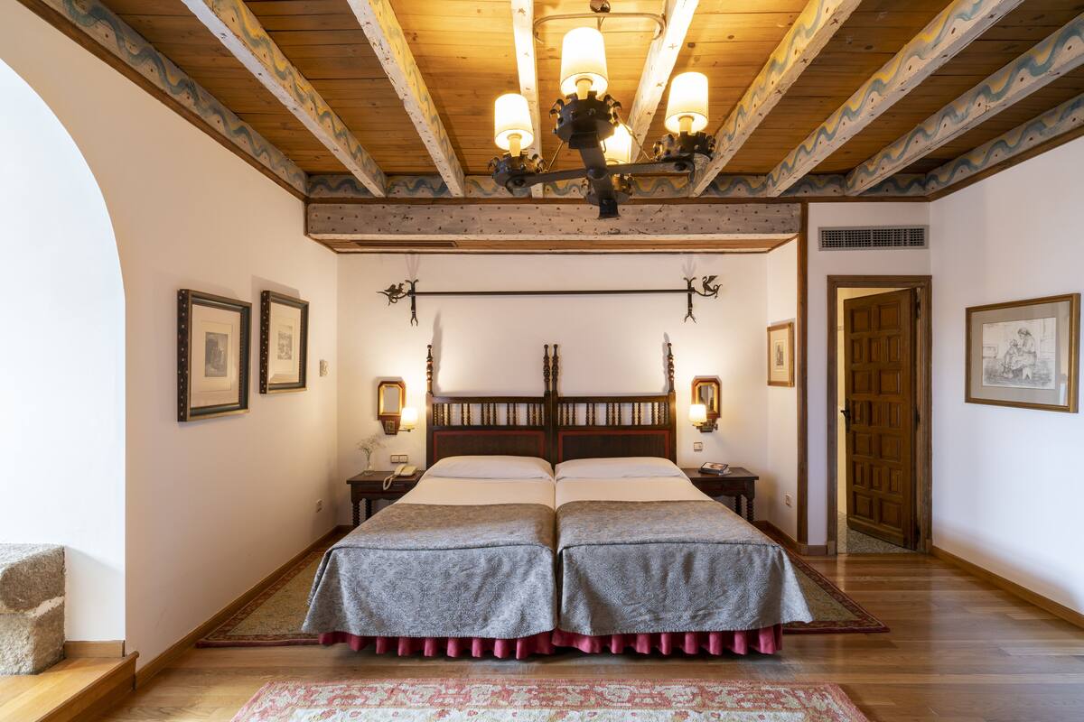 Quarto com duas camas, vigas de madeira, um candelabro, quadros emoldurados e um tapete estampado.
