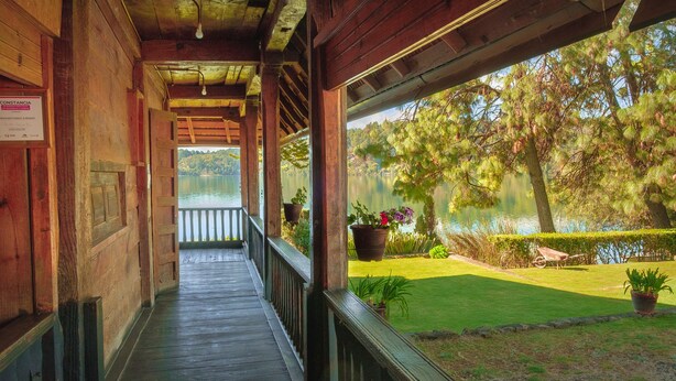A wooden covered walkway leading to a balcony overlooking a lake with potted plants and a lawn.