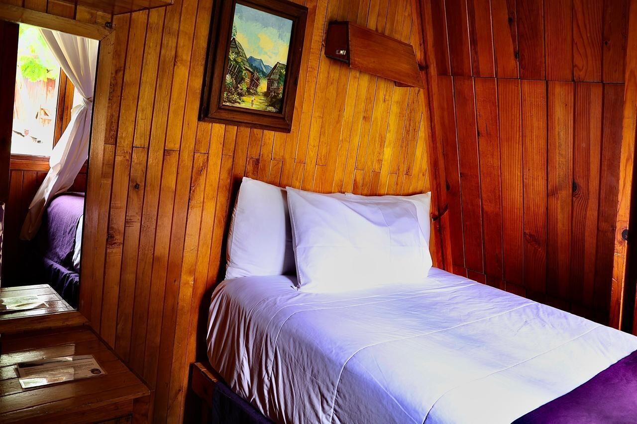 A wooden cabin room with a bed, a painting on the wall, and a window with curtains.