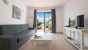 A modern living room with a grey sofa, a wooden coffee table, a flat-screen TV on a white stand, and a view through a glass door to an outdoor area with plants and a clear sky. - Apartamentos Santa Rosa (Teguise)