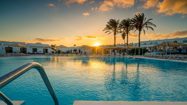 A swimming pool with lounge chairs and umbrellas, surrounded by palm trees and buildings during sunset. - Apartamentos Santa Rosa (Teguise)