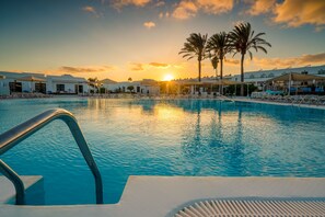 A swimming pool with lounge chairs and umbrellas, surrounded by palm trees and buildings during sunset.