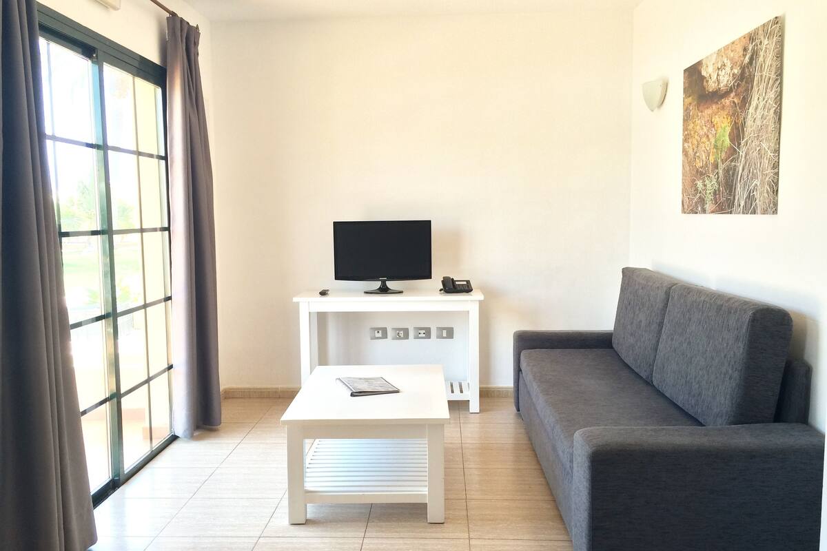 Una sala de estar moderna con un sofá gris, una mesa de centro blanca y un televisor sobre un mueble blanco.