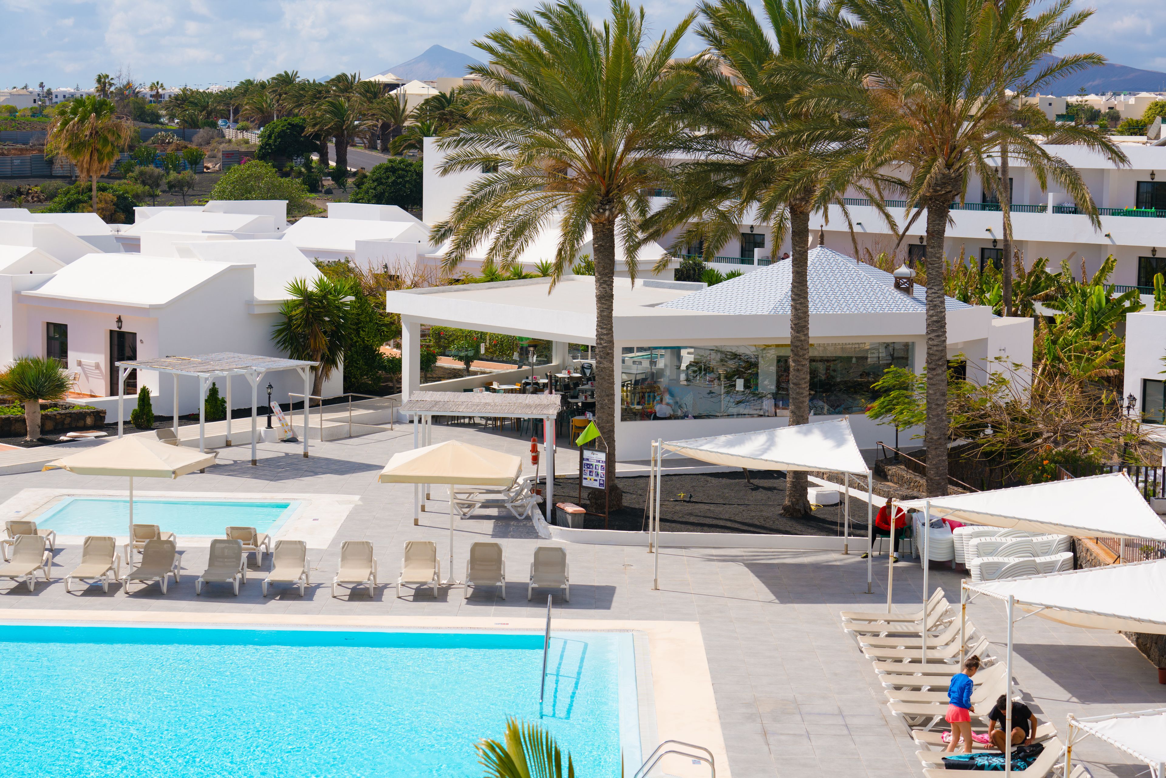 Un área de piscina con sillones reclinables y sombrillas, rodeada de edificios blancos y palmeras.