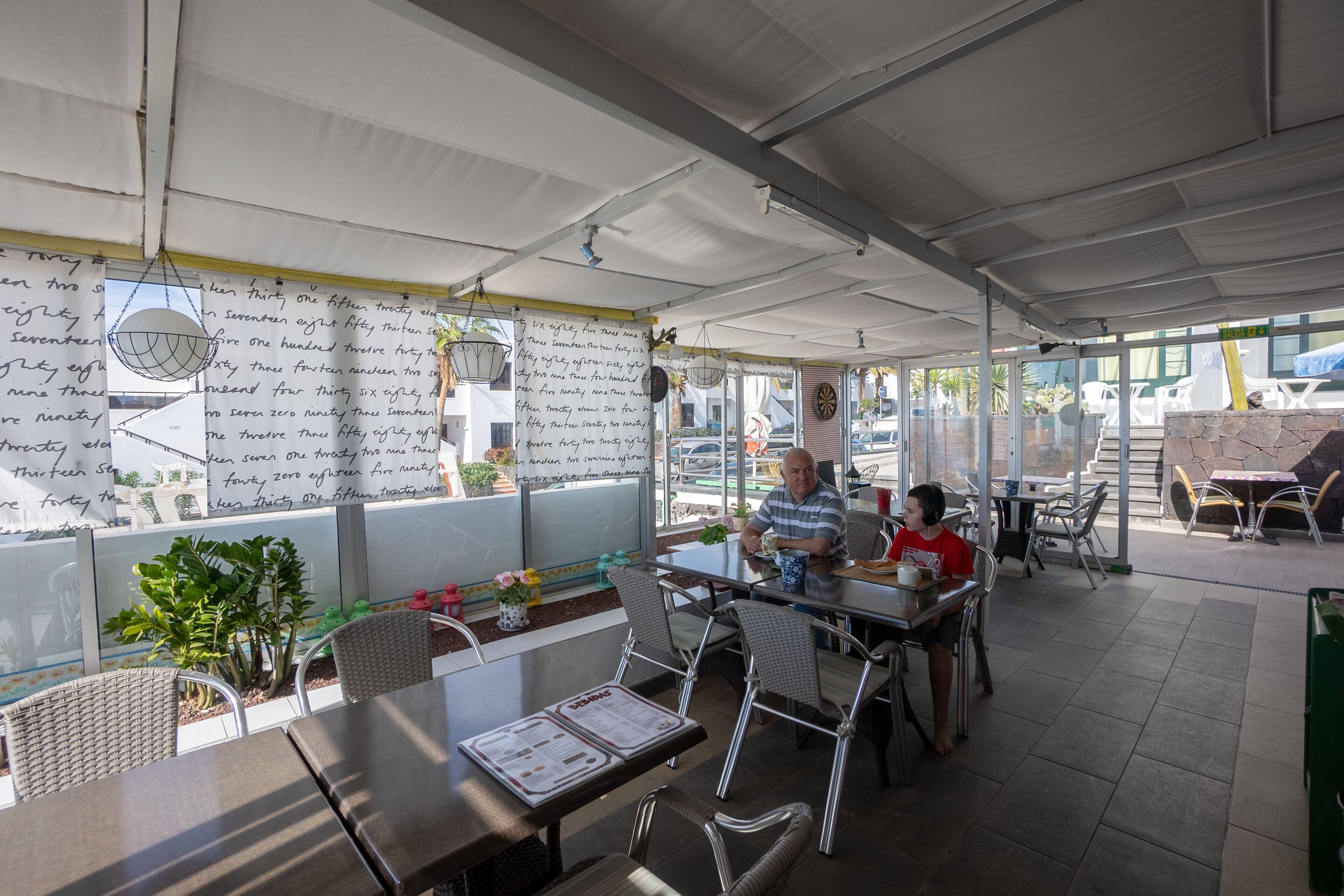 Un café en plein air avec des tables et des chaises, un menu et un mur où sont affichées des notes manuscrites.